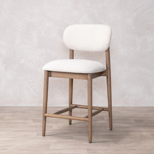 Bar stool with cream cushioned seat and backrest on a wooden floor against a light gray wall.