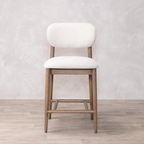 cream cushioned bar stool with wooden legs on a light wood floor against a light gray wall.
