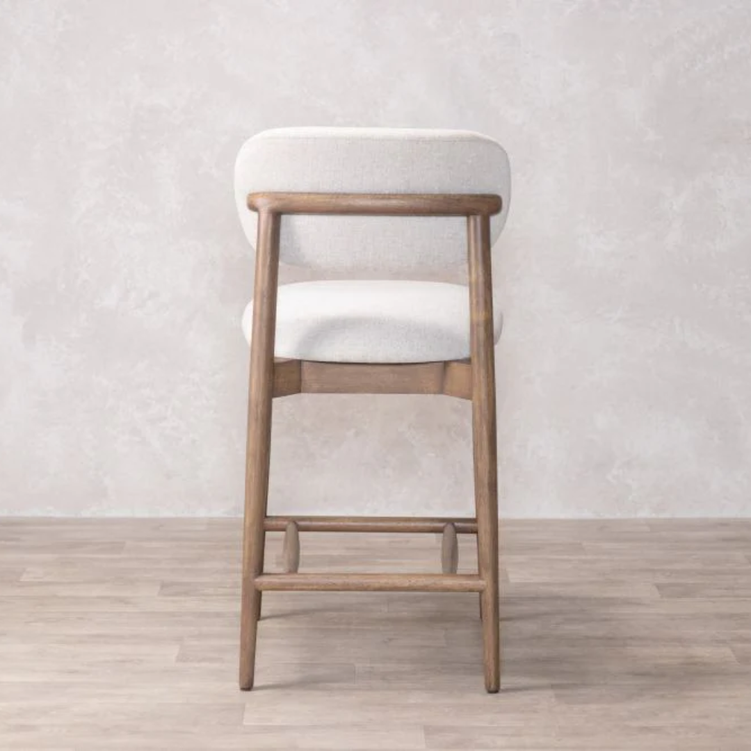Bar stool with wooden frame and cream  cushioned seat against a light gray wall.