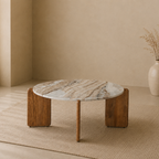 A round low coffee table with a white and gray marbled top and two thick wooden block legs, styled in a minimalist beige room on a woven rug.
