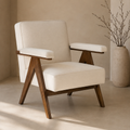A modern accent chair with cream-colored upholstered cushions and a dark brown wooden frame, set in a minimalist room with a beige wall and floor, next to a large textured vase with dried branches.