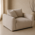 A beige upholstered armchair with two matching back cushions, shown in a softly lit neutral interior with a vase of branches in the background.