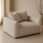 A beige upholstered armchair with two matching back cushions, shown in a softly lit neutral interior with a vase of branches in the background.