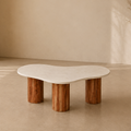 An organic-shaped coffee table with a smooth, wavy off-white stone top and three rustic, cylindrical brown wooden legs, set in a minimalist, warm-toned room.