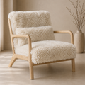 A modern wooden armchair with plush cream sheepskin-style upholstery, shown in a softly lit neutral interior with a beige wall and a vase of branches in the background.