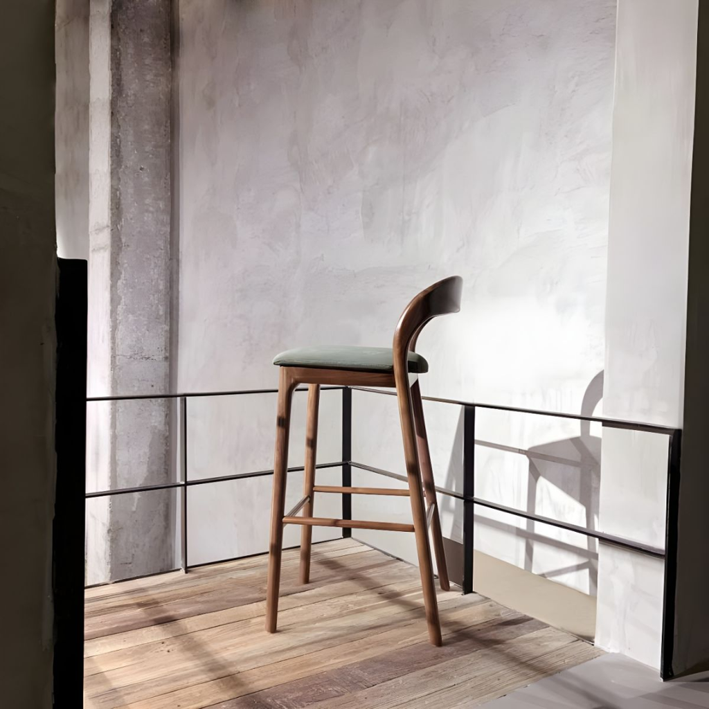 Wooden bar stool with a green leather seat on a wooden floor with a concrete wall in the background.