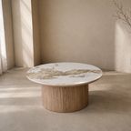 A round coffee table with a white marble top featuring gold and gray veining, supported by a fluted light oak wood base, set in a minimalist beige room.
