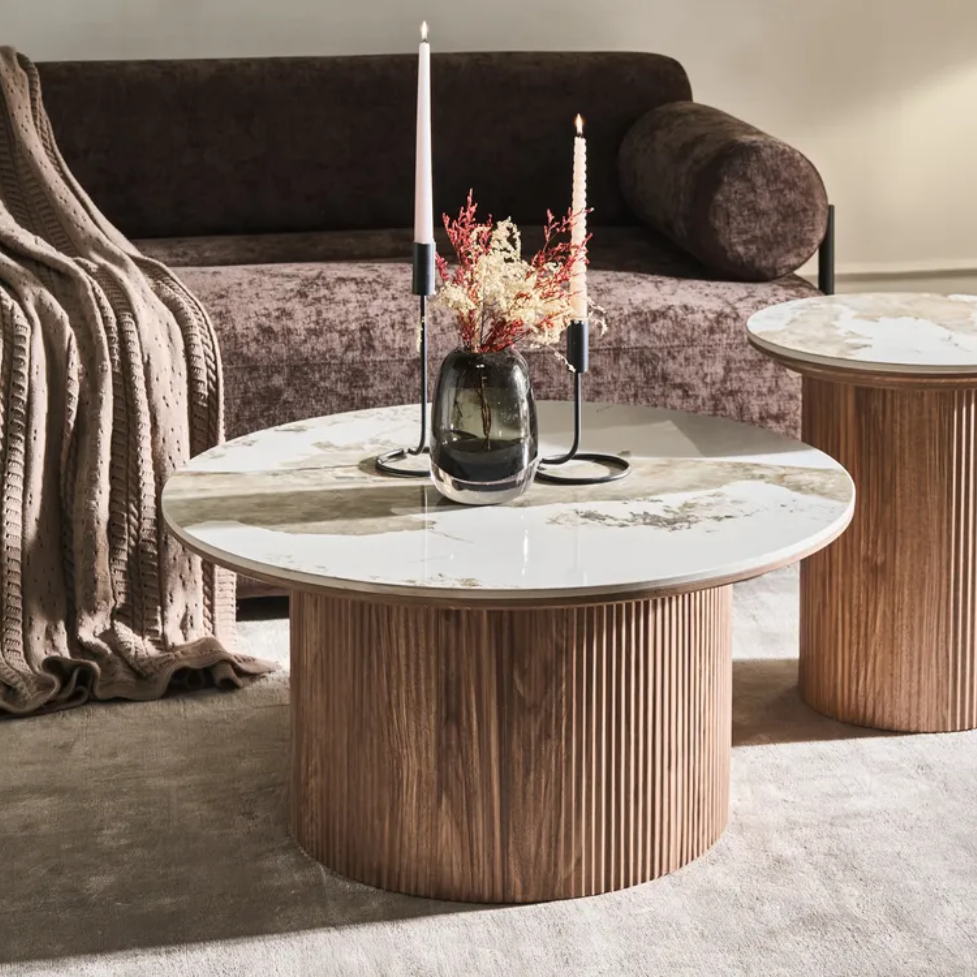 A modern round coffee table with a fluted wooden base and a white marble-patterned top, styled in a living room setting with a matching side table, a sofa, and a vase of dried flowers.