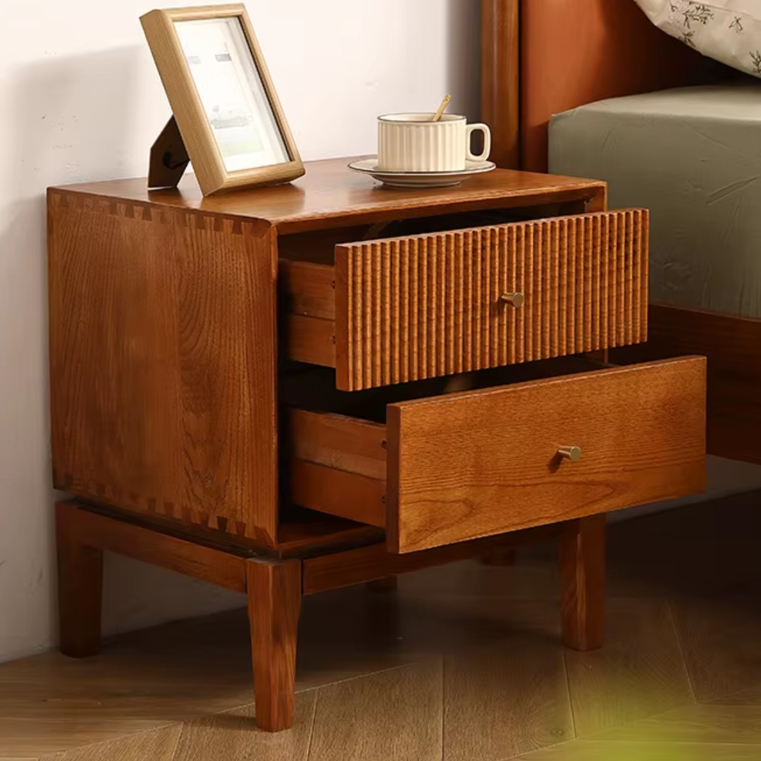 Wooden nightstand with two drawers, a cup, and a photo frame on top next to a bed.