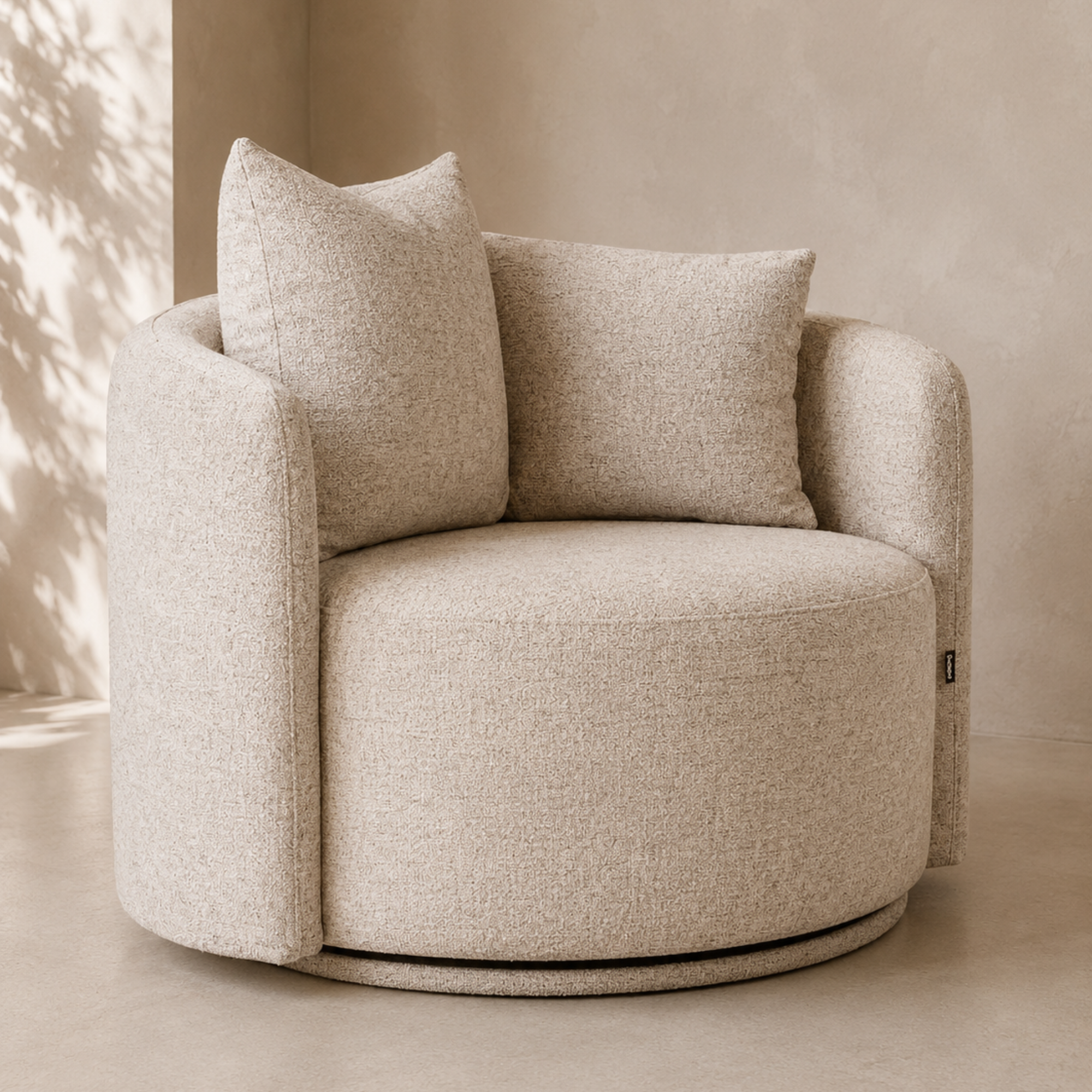 A round upholstered accent chair in a warm beige textured fabric, featuring two matching back cushions and a low circular base, photographed in a softly lit neutral interior.