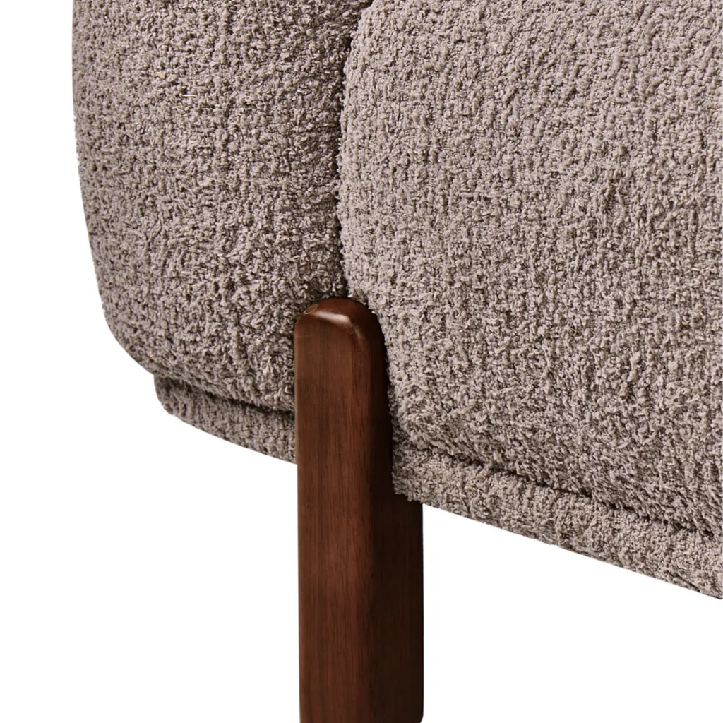 Close-up of a textured bouclé upholstered chair or sofa in a warm taupe-beige color, showing the fabric texture and a dark brown wooden leg against a white background.