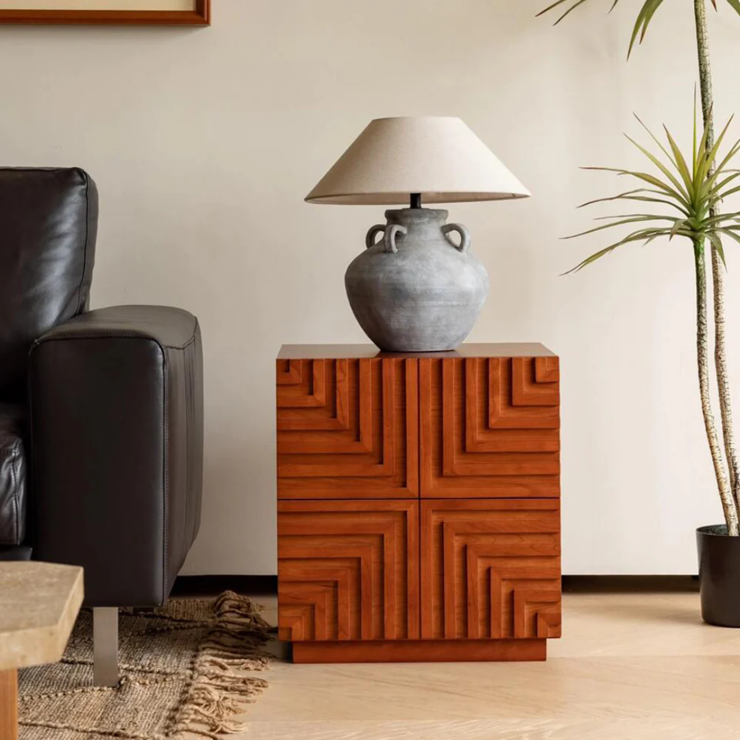 Wooden side table with geometric design, lamp, and plant in a living room setting.
