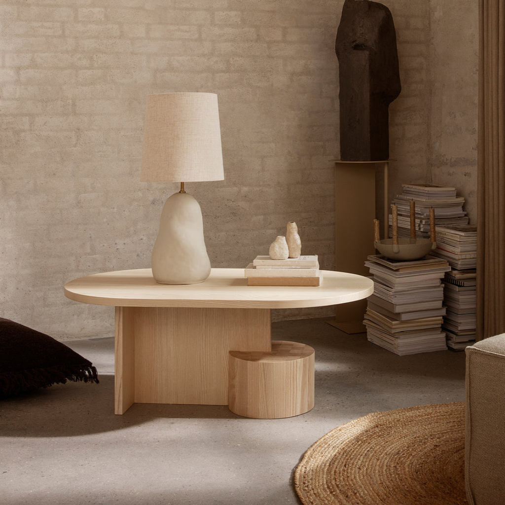  light Wood Oval Coffee Table With Sculptural Base with a lamp and decorative items against a textured wall.