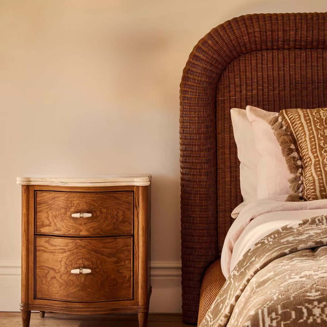 Walnut Bedside Table With Marble Top and Drawers next to a bed with wicker headboard and patterned bedding.